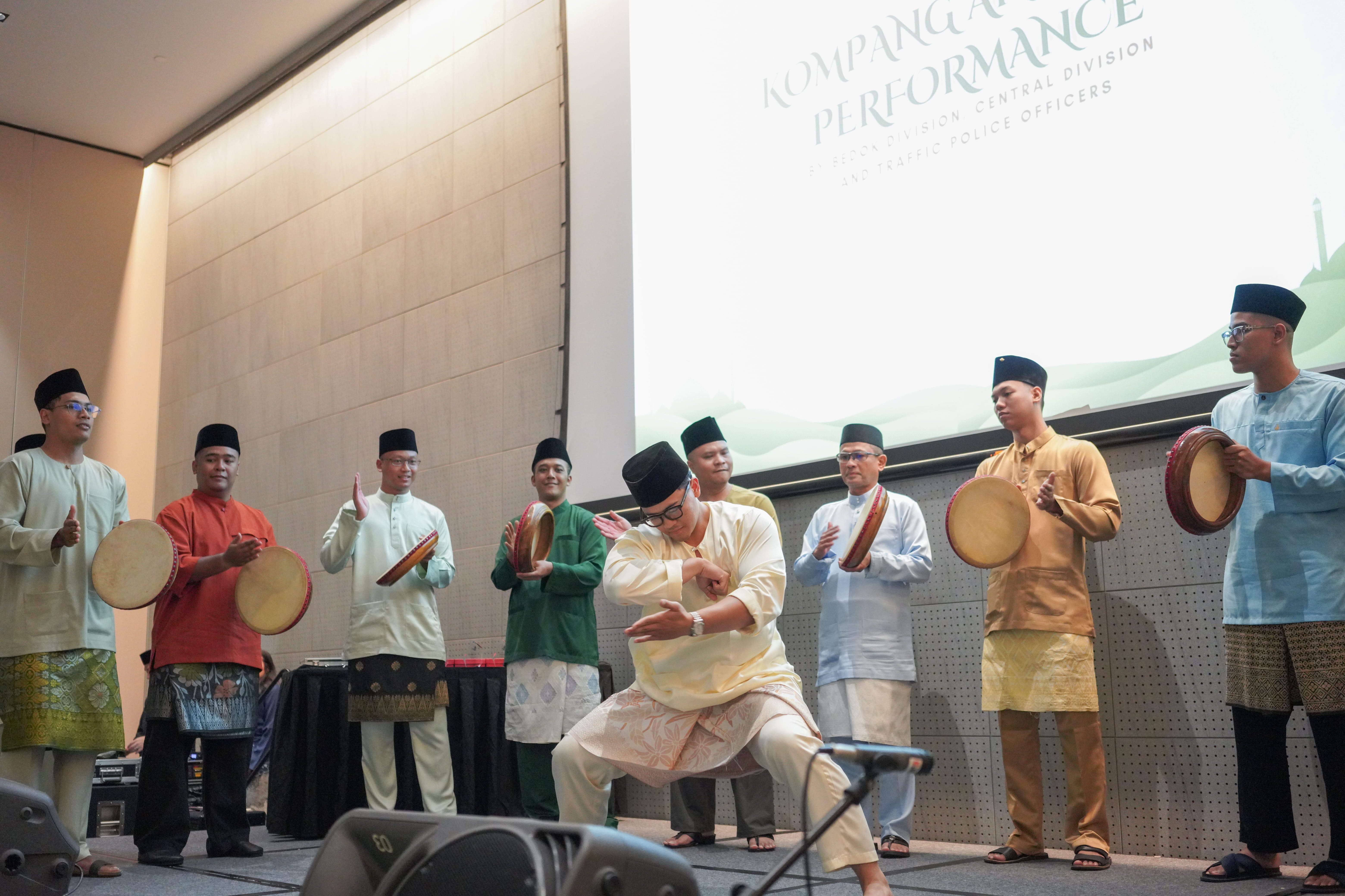 A silat performer striking a martial arts pose at centre stage while kompang musicians play frame drums around him.